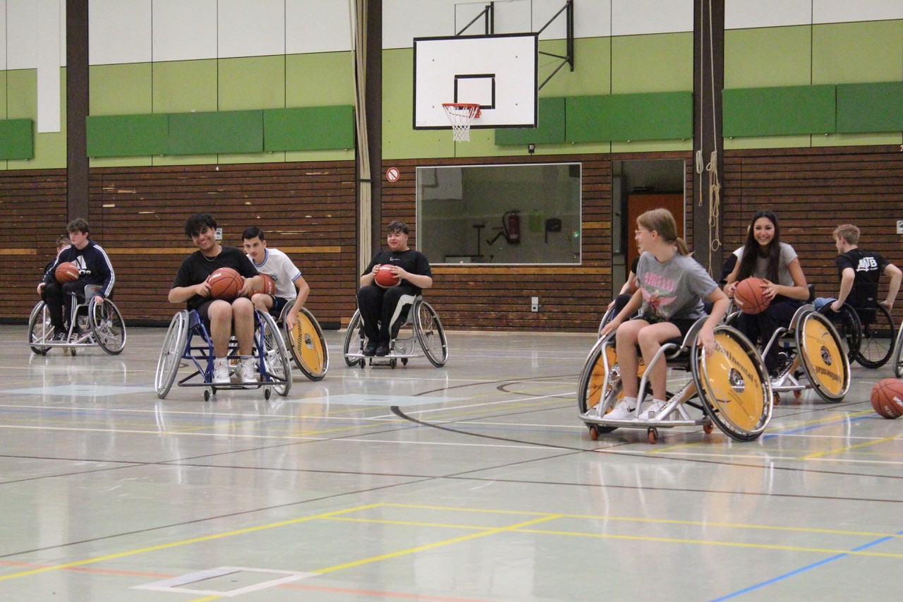 Mehrere Schülerinnen und Schüler spielen Rollstuhlbasketball in einer Sporthalle.