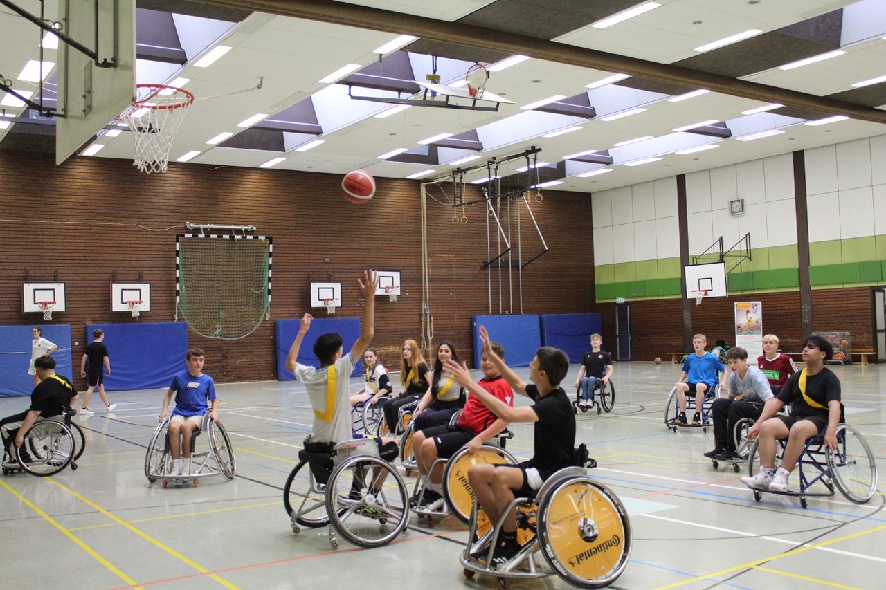 Mehrere Schülerinnen und Schüler spielen Rollstuhlbasketball in einer Sporthalle.